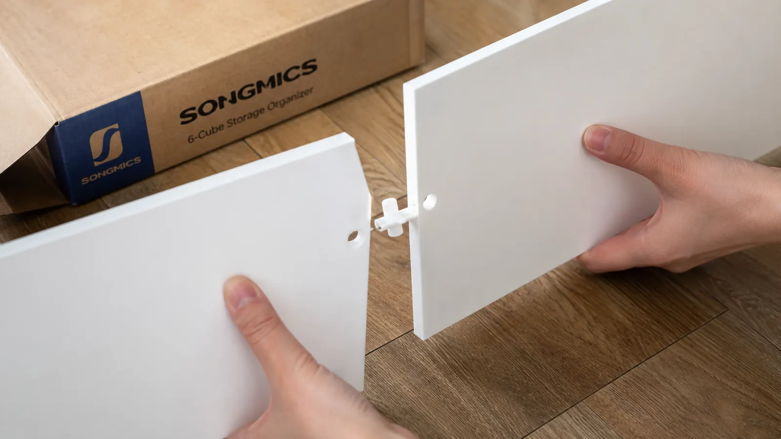 Hands assembling a white modular cube shelf panel by snapping connectors together on a wood floor