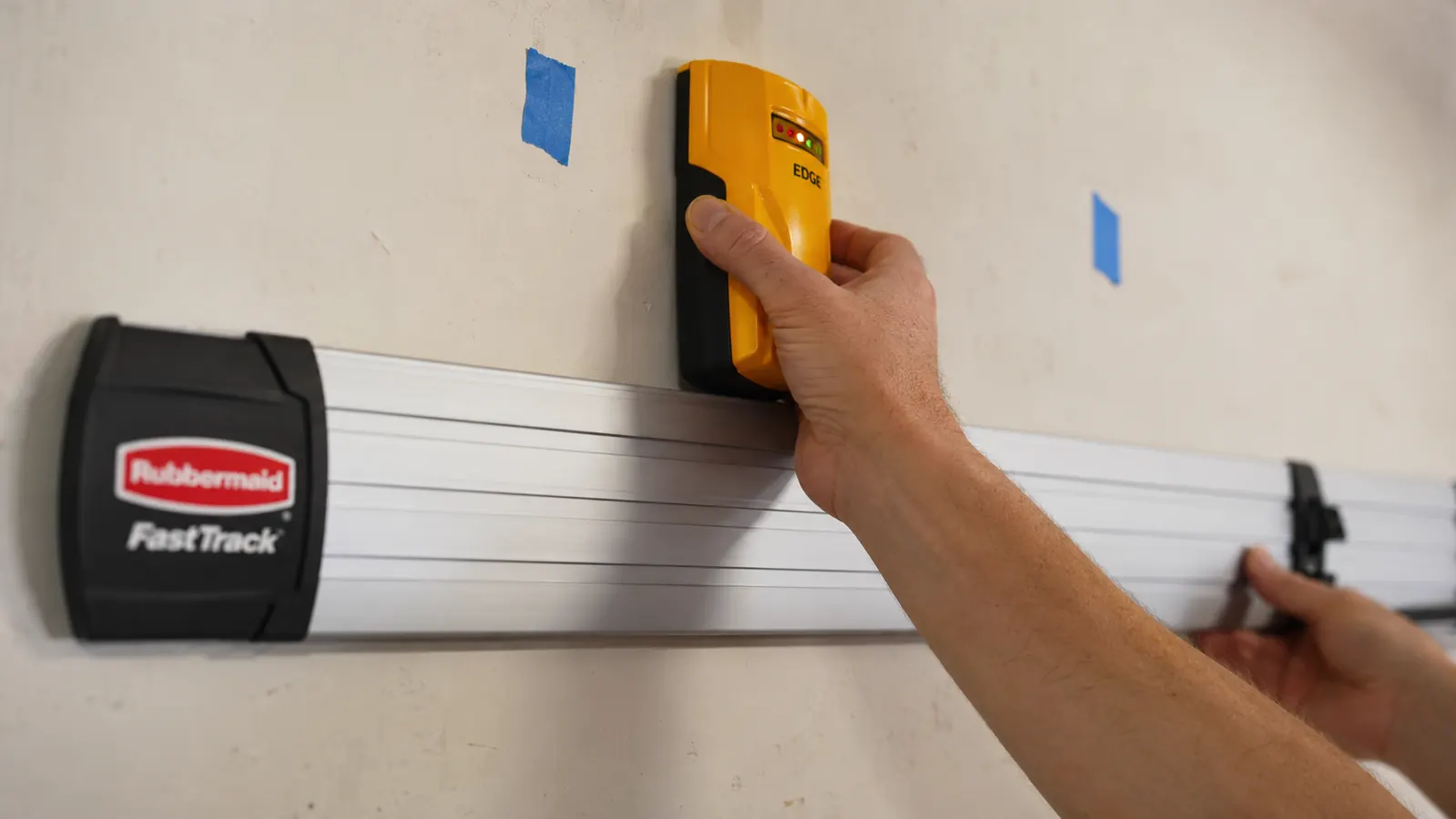 Hand holding a stud finder against a garage drywall wall while marking stud locations with painter's tape