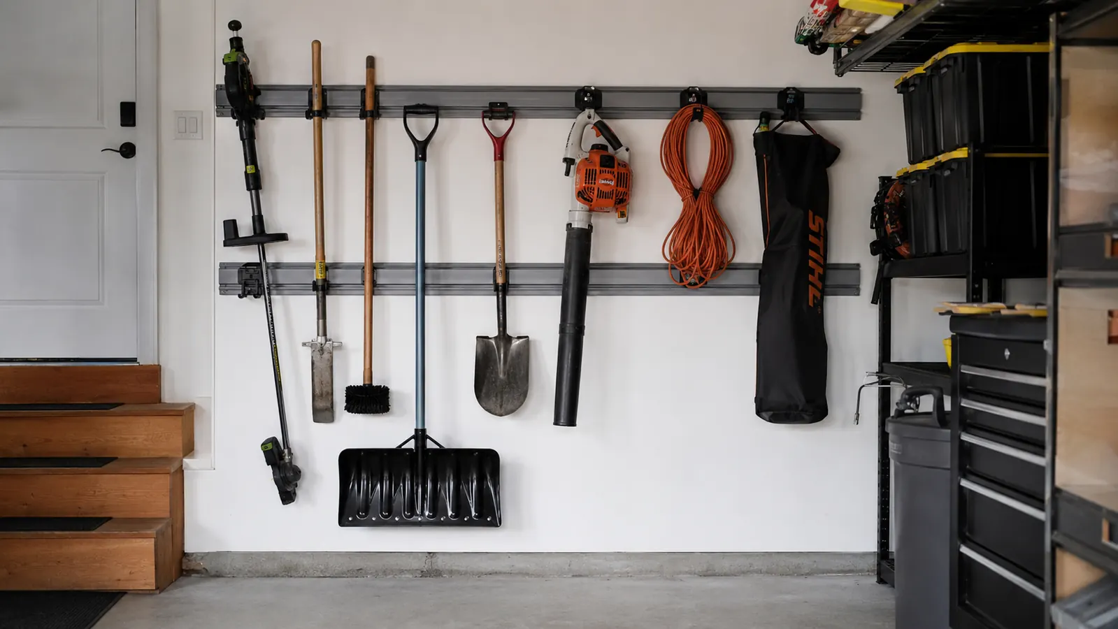 Garage wall corner with Rubbermaid FastTrack rails holding a snow shovel, leaf blower, and extension cord hook in an organized arrangement